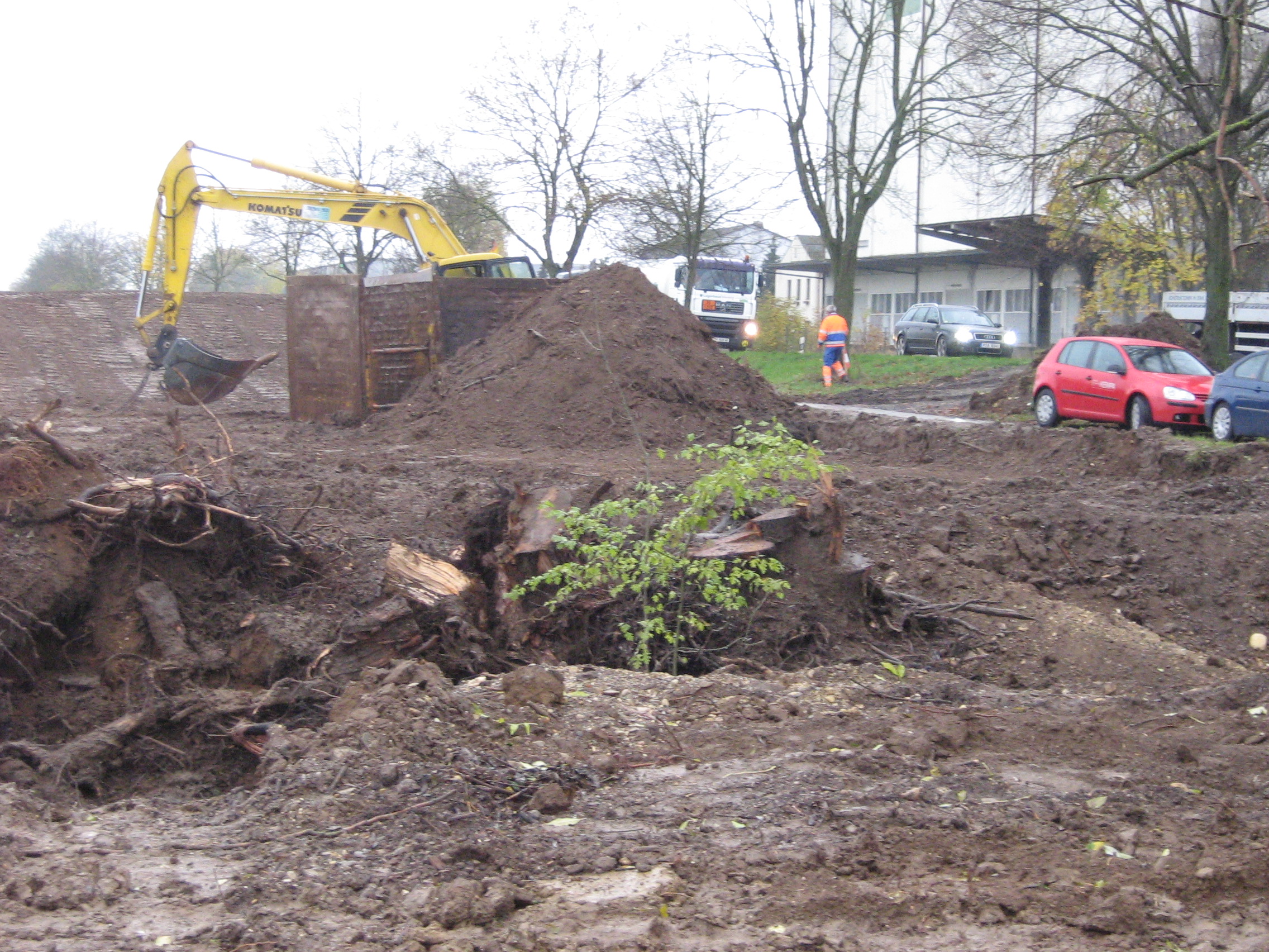 Vor dem Baubeginn wird das Baufeld von Sträucher und Bäumen samt Wurzeln freigeräumt. Ein Baggerarm ist in der Mittes des Bildes zu sehen. Dieser steht hinter einem Erdhaufen und Wänden zur Baugrubensicherung. Ringsrum sind Erdreich, Wurzeln, Baumreste. Im Hintergrund rechts die Straße mit Fahrzeugen und ein Baurabeiter. 